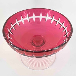 Puede incluir: Un cuenco vintage de cristal de color rojo oscuro sobre un pedestal de cristal transparente. El cuenco tiene un dise&ntilde;o de cristal tallado alrededor del borde y un borde festoneado.