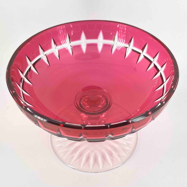 Puede incluir: Un cuenco vintage de cristal de color rojo oscuro sobre un pedestal de cristal transparente. El cuenco tiene un dise&ntilde;o de cristal tallado alrededor del borde y un borde festoneado.