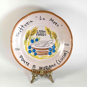 May include: A round ceramic plate with a light cream glaze and a brown rim. The center features a hand-painted illustration of a bowl with a spoon, wheat stalks, and blue berries. Text reads: Trattoria Lu Mosa, Gran Farro, 1964, 1984, Ponte a Moriano (Lucca).