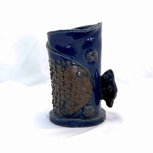 May include: A ceramic vase in the shape of a fish, with a dark blue glaze. The fish has a brown textured body and a black tail fin. Seashells are also featured on the vase.