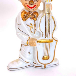 Puede incluir: Figurilla de porcelana blanca de un payaso tocando un violonchelo. El payaso lleva un traje blanco con una pajarita dorada y tiene la nariz roja y los labios pintados de rojo. El violonchelo est&aacute; pintado de oro con cuerdas negras.