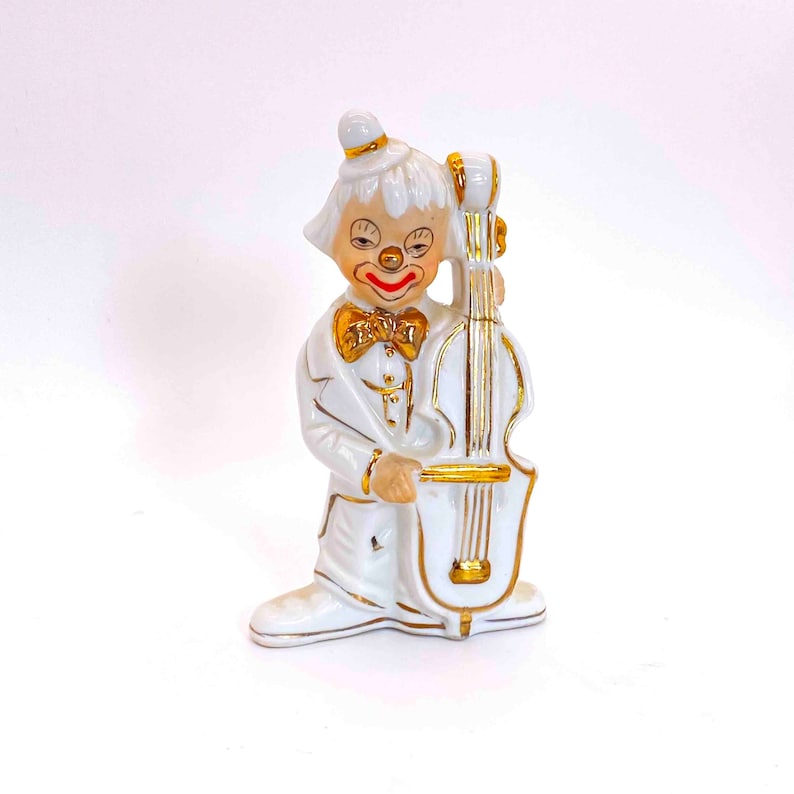 Puede incluir: Figurilla de porcelana blanca de un payaso tocando un violonchelo. El payaso lleva un traje blanco con ribetes dorados y una pajarita roja. El violonchelo tambi&eacute;n es blanco con ribetes dorados.