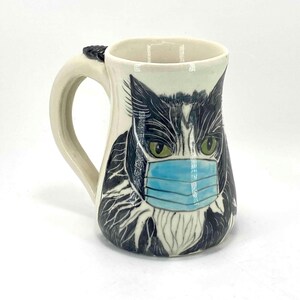 May include: A white ceramic mug with a black and white cat wearing a blue face mask. The cat's eyes are green and the mug has a brown handle.