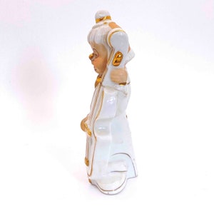 Puede incluir: Figurita de porcelana blanca de una persona tocando un violonchelo. La figurita est&aacute; decorada con detalles dorados.