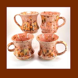 May include: Set of four handmade ceramic mugs with a pink and yellow speckled glaze. The mugs have a wide base and a narrow top, with a rounded handle.