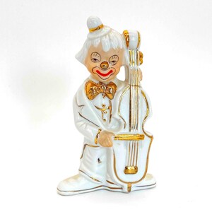 Puede incluir: Figurilla de porcelana blanca de un payaso tocando un violonchelo. El payaso lleva un traje blanco con una pajarita dorada y un sombrero blanco. El violonchelo est&aacute; decorado con detalles dorados.
