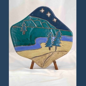 May include: A ceramic wall hanging with a blue night sky, white stars, green mountains, a blue river, and three green trees. The wall hanging is on a wooden stand.