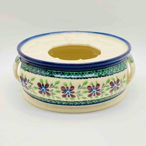 Può includere: Un vaso in ceramica bianca con disegni floreali blu e verdi e due manici. Il vaso ha una sezione centrale rialzata con un foro al centro.