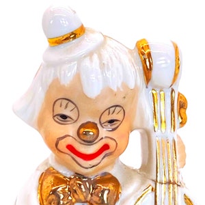 Puede incluir: Una figurita de porcelana blanca de un payaso tocando el viol&iacute;n. El payaso lleva un sombrero blanco con una banda dorada, una pajarita blanca y una sonrisa roja. El viol&iacute;n est&aacute; decorado con detalles dorados.