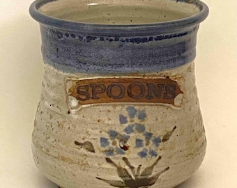 Puede incluir: Un porta cucharas de cerámica con cuerpo color crema moteado y borde azul oscuro. El frente presenta una placa rectangular en relieve con la palabra SPOONS grabada en marrón y un diseño floral pintado en azul y verde bajo la etiqueta.