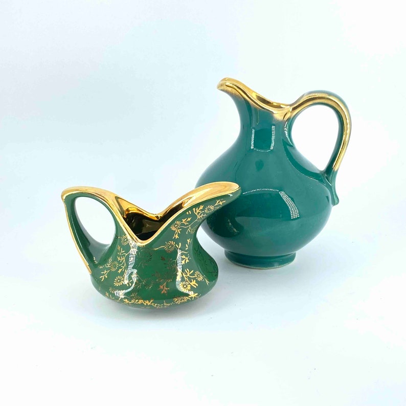 Puede incluir: Dos jarras de cer&aacute;mica verde con detalles dorados. La jarra m&aacute;s peque&ntilde;a tiene un dise&ntilde;o floral decorativo. La jarra m&aacute;s grande tiene un dise&ntilde;o simple.