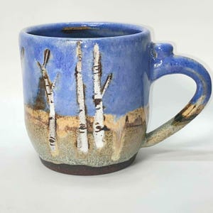 Puede incluir: Taza de cerámica hecha a mano con un diseño de paisaje. La taza presenta un cielo azul, suelo marrón y abedules blancos. El asa y el borde tienen un esmalte azul. Una pieza única de arte funcional.