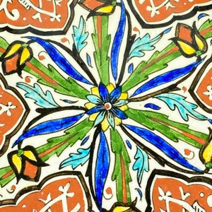 Puede incluir: Un primer plano de un azulejo de cerámica con un diseño floral colorido. El azulejo presenta una flor central con pétalos azules, amarillos y blancos, rodeada de hojas verdes y acentos azules y rojos.