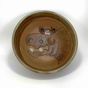 Può includere: Una ciotola in ceramica marrone con un viso di cartone animato fantasioso scolpito all'interno. Il viso ha occhi e denti bianchi e un naso marrone.