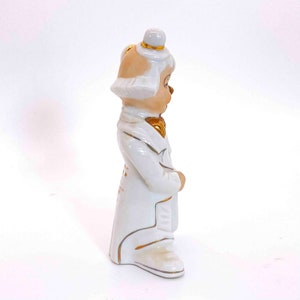 Puede incluir: Una figurita de porcelana blanca de un payaso que lleva un traje blanco con detalles dorados. El payaso lleva un sombrero blanco con una banda dorada y una pajarita dorada. La figurita est&aacute; de pie sobre un fondo blanco.