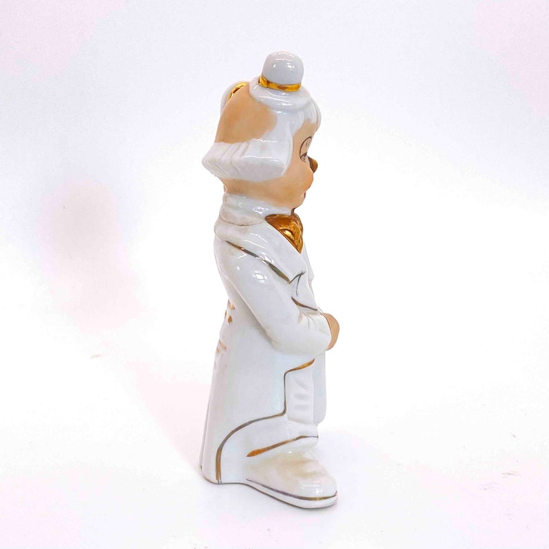Puede incluir: Una figurita de porcelana blanca de un payaso que lleva un traje blanco con detalles dorados. El payaso lleva un sombrero blanco con una banda dorada y una pajarita dorada. La figurita est&aacute; de pie sobre un fondo blanco.
