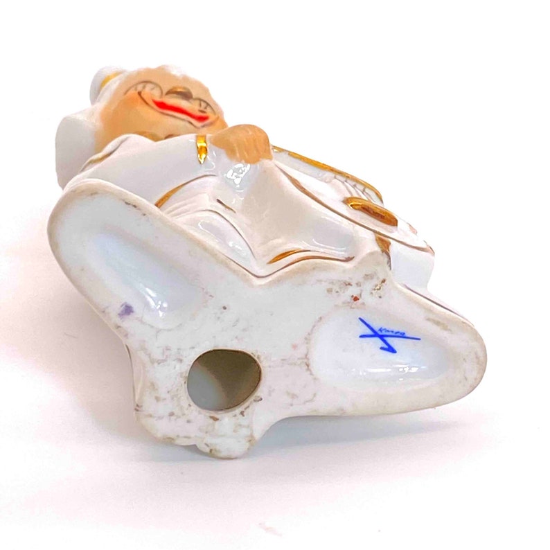 Puede incluir: Figurita de porcelana blanca de una persona tocando un instrumento musical. La figurita est&aacute; decorada con pintura dorada y tiene una marca azul en la parte inferior.