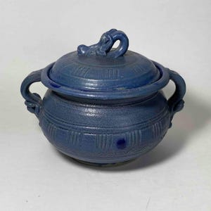 Pode incluir: Uma tigela redonda de cerâmica azul cobalto com tampa, apresentando duas alças laterais e uma pega esculpida em forma de animal na tampa. A superfície exterior é decorada com padrões de linhas verticais e uma base texturizada, com acabamento em verniz brilhante.