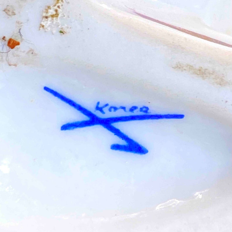 Puede incluir: Firma manuscrita azul con la palabra "Korea" sobre una superficie de porcelana blanca.