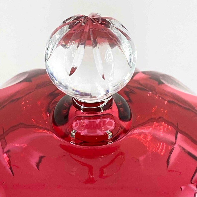 Puede incluir: Un pomo de vidrio transparente con un dise&ntilde;o facetado se encuentra encima de una tapa de vidrio roja. La tapa es probablemente para un frasco o recipiente decorativo.
