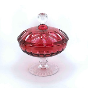 Puede incluir: Un platito de cristal transparente con un cuenco rojo facetado y una tapa de cristal transparente con un pomo. El plato se asienta sobre un pedestal de cristal transparente.