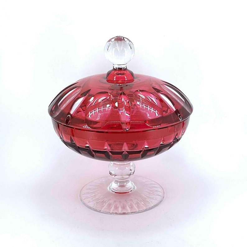 Puede incluir: Un platito de cristal transparente con un cuenco rojo facetado y una tapa de cristal transparente con un pomo. El plato se asienta sobre un pedestal de cristal transparente.