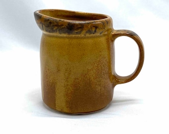 Puede incluir: Una jarra de crema de cerámica marrón con un borde marrón y negro y un asa. La jarra tiene un esmalte moteado.