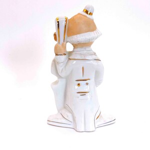 Puede incluir: Figurilla de porcelana blanca de un payaso sosteniendo un instrumento musical. El payaso lleva un traje blanco con ribetes dorados y un sombrero blanco. La figurita mide aproximadamente 15 cm de alto.