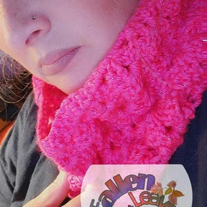 May include: A bright pink cowl crochet pattern with silver accents. The cowl is made of a chunky yarn and is wrapped around the neck. The image includes the text "Fallen Leaves" and "Cowl Crochet Pattern".