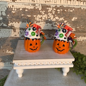 May include: Two Halloween-themed pumpkin mugs filled with candy, including Skittles, M&Ms, and candy eyeballs. The mugs are sitting on a white pedestal with a beaded trim.