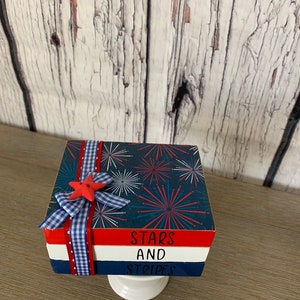 May include: A stack of three wooden blocks with a red, white, and blue patriotic theme. The top block has a blue background with white fireworks and red accents. The middle block is red with white text that says "Stars". The bottom block is white with blue text that says "And Stripes". The blocks are tied together with a blue and white gingham ribbon and a red star.