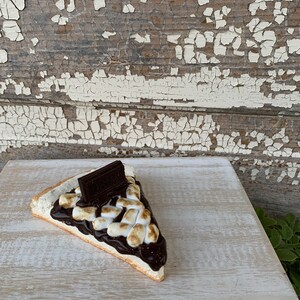 May include: A slice of s'mores pie with a chocolate bar on top. The pie is on a white wooden surface with a white base.