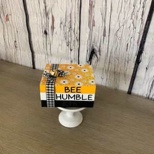 May include: A decorative wooden block with the words "BEE HUMBLE KIND" in black and white. The block is yellow with white daisy patterns and a black and white checkered ribbon. A small bee embellishment is attached to the ribbon. The block sits on a white pedestal.