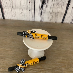May include: Two miniature yellow rolling pins with black handles and black and white checkered bows. Each rolling pin has the words "Bee Kind" written on it.