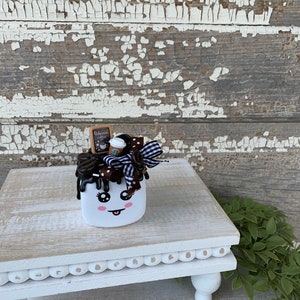May include: A whimsical white marshmallow-shaped decoration with a smiling face, chocolate drizzle, and a coffee cup. It is adorned with a black and white bow and a small sign that reads "Delicious Coffee". The decoration sits on a white, distressed wooden stand.
