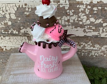 May include: A pink ceramic ice cream cone shaped watering can with a brown and white ice cream cone on top. The watering can has the text "Dairy Fresh Ice Cream" on the side.