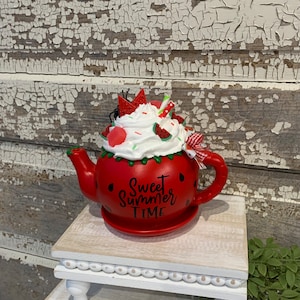 May include: A red teapot with a watermelon design, topped with whipped cream and watermelon slices. The teapot has a handle and spout, and the words "Sweet Summer Time" are written on the front. It sits on a small, white, decorative stand.