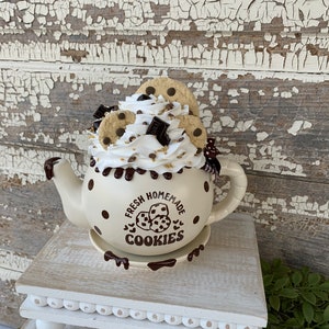 May include: A white ceramic teapot with brown polka dots and a chocolate glaze. The teapot is filled with a white whipped cream topping with chocolate chips and cookie pieces. The teapot has the text "Fresh Homemade Cookies" on the side.
