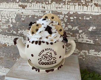 May include: A white ceramic teapot with brown polka dots and a chocolate glaze. The teapot is filled with a white whipped cream topping with chocolate chips and cookie pieces. The teapot has the text "Fresh Homemade Cookies" on the side.