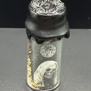 May include: A small glass vial with a black cap and seal. The vial features a graphic of a hooded skull with the word "DEATH" below it. The cap has a decorative silver design.