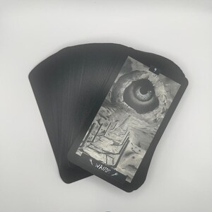 May include: A deck of black tarot cards with a grayscale image of an eye inside a cave on the front of one card. The card reads "9" and "Wands". The cards are fanned out on a white surface.