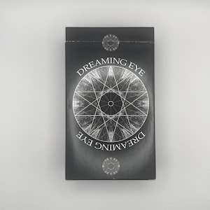 May include: A black rectangular box with the words "Dreaming Eye" in white lettering. A silver geometric design is centered on the front. Two silver circular designs are at the top and bottom of the box.