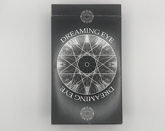 Op de afbeelding: Een zwarte rechthoekige doos met de woorden "Dreaming Eye" in witte letters. Een zilverkleurig geometrisch ontwerp is gecentreerd op de voorkant. Twee zilverkleurige cirkelvormige ontwerpen bevinden zich aan de boven- en onderkant van de doos.