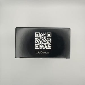 May include: A black box with a QR code and the text "L.A.Duncan" printed in white. The box is rectangular and appears to be made of cardboard or a similar material. The background is a light gray color.
