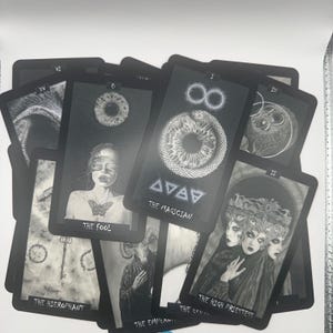 May include: A deck of tarot cards with black borders and grayscale illustrations. The cards feature images of The Fool, The Magician, The High Priestess, and The Hierophant. The deck has a gothic or mystical theme.