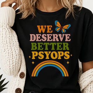 May include: Black t-shirt with the text "WE DESERVE BETTER PSYOPs" in a colourful retro font. A butterfly graphic and rainbow design are also featured. The shirt is a casual style.