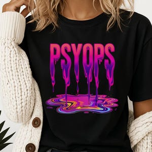 May include: Black t-shirt with the word "PSYOP" in a gradient pink and purple dripping design. The dripping design pools at the bottom in a colorful, abstract shape. The shirt is a crew neck style.