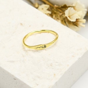 Puede incluir: Un anillo dorado con un diseño delicado. El anillo presenta una banda delgada y una cara rectangular con un patrón floral grabado y una pequeña piedra negra. El anillo se exhibe sobre una superficie blanca texturizada con flores secas en el fondo.