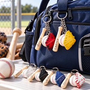 Puede incluir: Una colección de llaveros de béisbol con bates de madera en miniatura grabados con nombres como Bruce, Carter, Harley y Arthur, junto a pequeñas pelotas y guantes coloridos en rojo, amarillo, blanco, negro y azul, sujetos a un bolso azul marino.