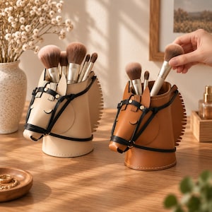 Puede incluir: Dos soportes cilíndricos para brochas con forma de cabeza de caballo, uno en color crema y otro en color canela. Cada uno presenta una brida negra, ojos cerrados y una crin en la parte posterior. Contienen varias brochas de maquillaje.